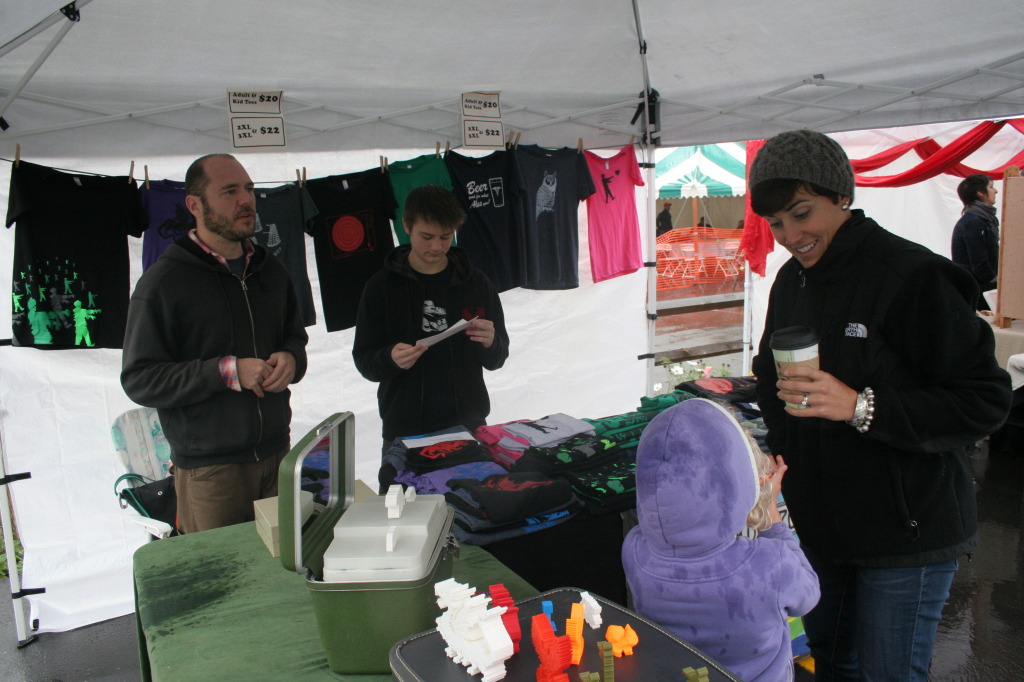 Despite rains and overcast skies, people still turned out to support the arts at the City of the Hills  Arts and Music Festival on Saturday where local artisans, musicians, and eateries had their creations on display. Here, at the Blackbird and Peacock tent, Joesph Von Stengal, Oneonta, and his son Soren Smeltzer chat with Corrinne Smith, Oneonta, and her daughter Claire, who saught shelter under the tent, but stayed for to look at the tee shirts and 3d-printed sculptures. (Ian Austin/ allotsego.com